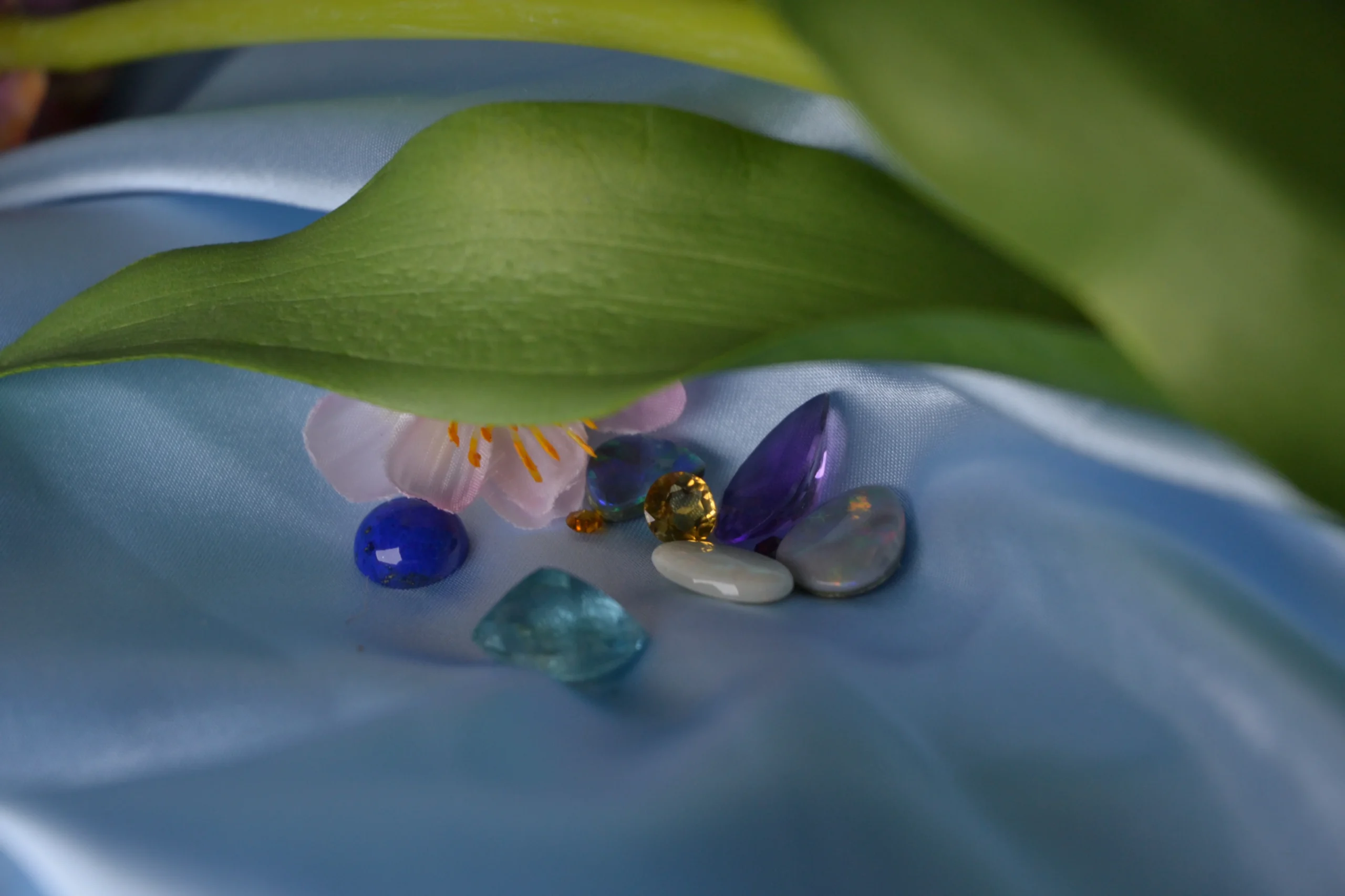 Loose colourful gemstones including opal, amethyst, and blue topaz ready for bespoke jewellery remodelling at Alexander Jewellery Chichester, displayed on blue silk.