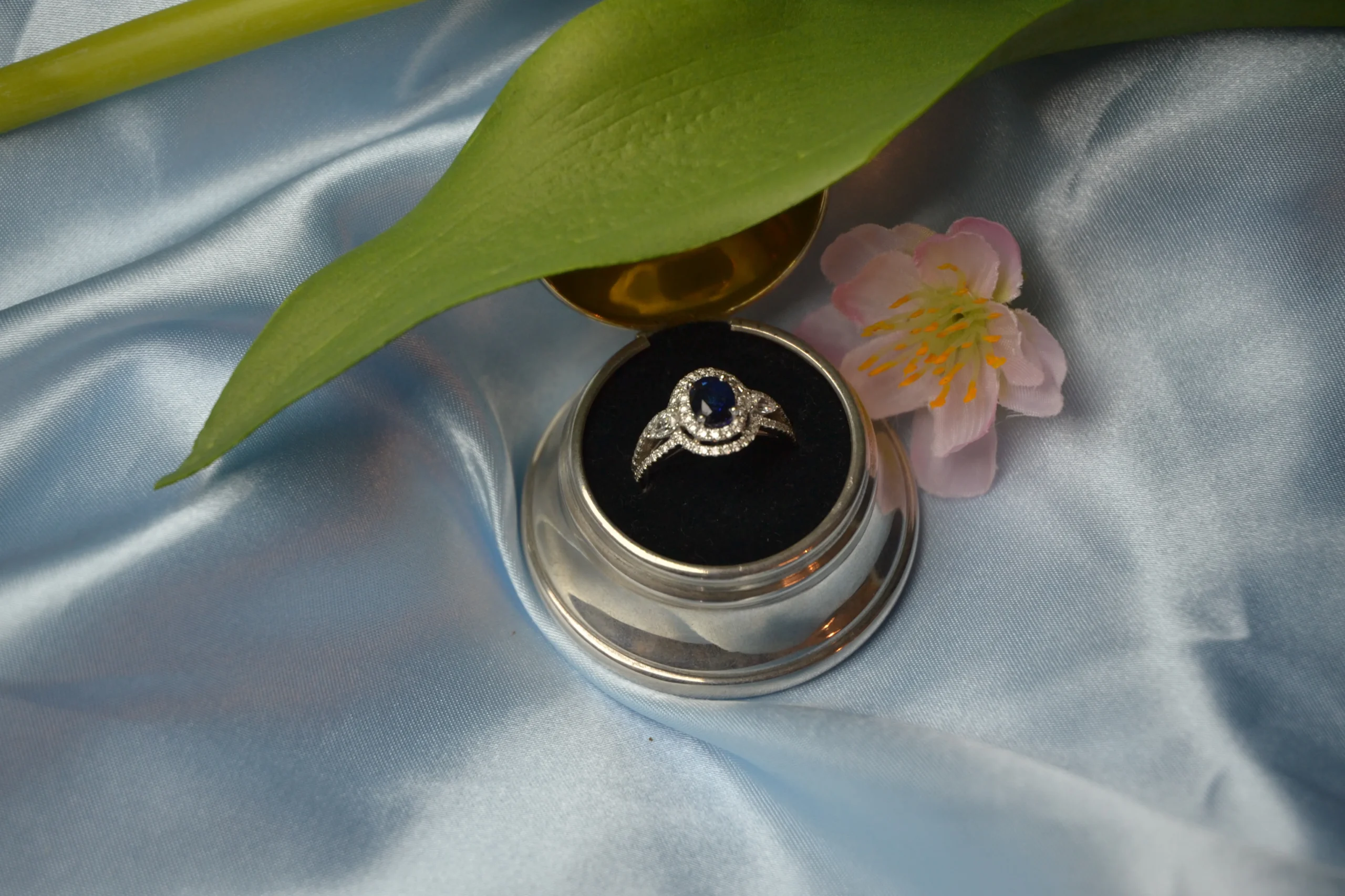 Bespoke sapphire and diamond halo engagement ring displayed in a round silver box on blue silk by Alexander Jewellery in Chichester.